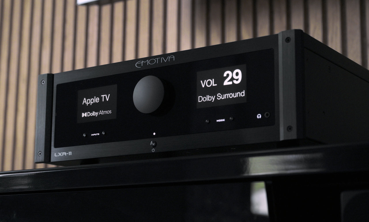 A black Emotiva LXR-11 AV receiver with a digital display screen, resting on a dark stand against a wooden slatted background.