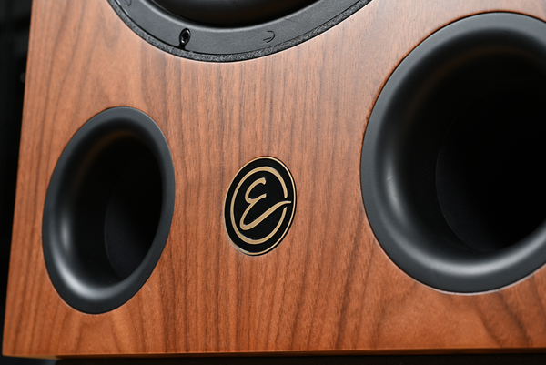 Close-up of a wooden speaker cabinet with black circular driver surrounds and a central oval logo badge featuring an 'E' monogram.