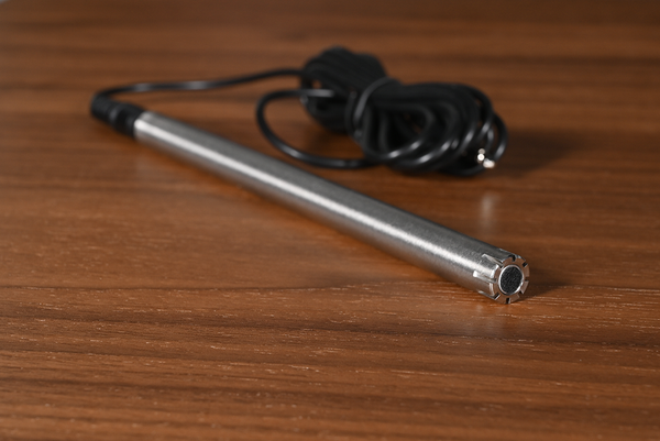 A stainless steel cylindrical measurement mic with a black connector and attached black power cord, resting on a brown wooden surface.