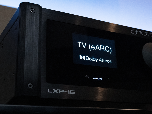 Close-up of a black LXP-16 audio receiver or amplifier with a lit display screen showing 'TV (eARC)' and 'Dolby Atmos'. The front panel has brushed metal details, screws, and a circular control knob partially visible on the right.