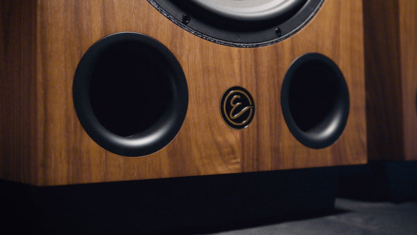 Wooden speaker with two black ports and a brand logo on a dark background